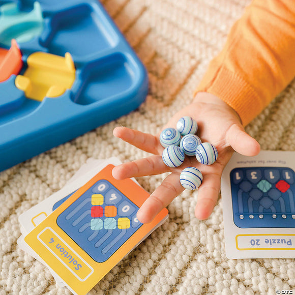 A child's hand holding colorful marbles, with a blue mechanical base and challenge cards in the background. The cards display different puzzle solutions. The setting features a light-colored woven rug.