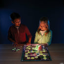 Two children, a boy and a girl, play the Ghost in the Attic game in a dimly lit room. The boy leans forward, focused on the game board which features a glowing green ghost piece and various character figures. The girl, smiling, points to the board while holding a flashlight, illuminating the spooky themed path. A spinner with ghostly graphics is placed on the side.