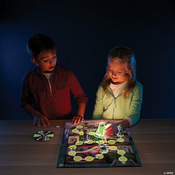Two children, a boy and a girl, play the Ghost in the Attic game in a dimly lit room. The boy leans forward, focused on the game board which features a glowing green ghost piece and various character figures. The girl, smiling, points to the board while holding a flashlight, illuminating the spooky themed path. A spinner with ghostly graphics is placed on the side.
