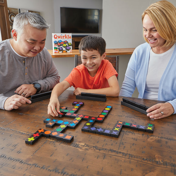 A family enjoying a game of Qwirkle, made by MindWare, featuring three players at a wooden table. The joyful boy in an orange shirt reaches for a colorful tile while his mother and father watch with smiles. The game involves colorful shapes on black tiles arranged in a playful pattern, promoting strategy and fun interactions.