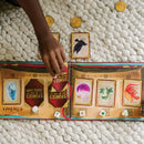 A hand reaches for a card from the board game 'Stories of the Three Coins' by Mindware. The game features several colorful cards representing various characters and items, displayed alongside golden coin tokens on a textured blanket.