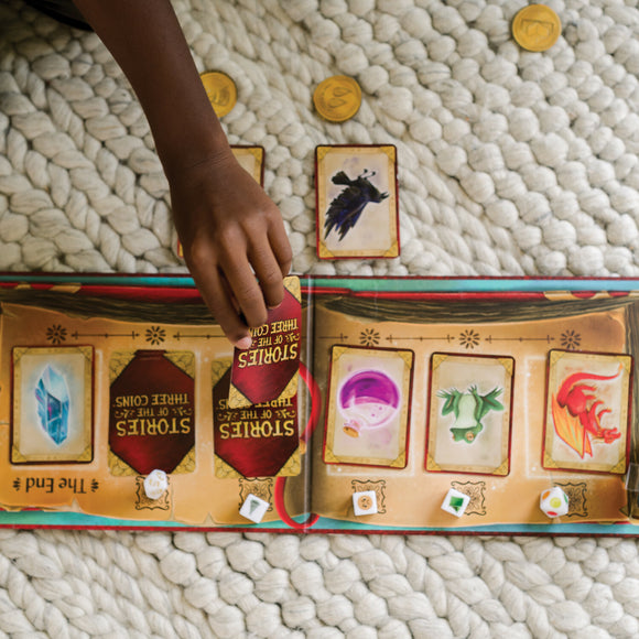 A hand reaches for a card from the board game 'Stories of the Three Coins' by Mindware. The game features several colorful cards representing various characters and items, displayed alongside golden coin tokens on a textured blanket.