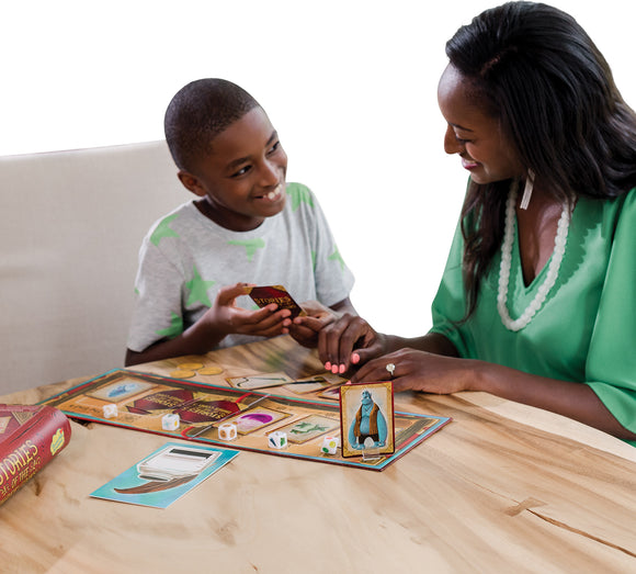 A child and an adult woman are engaged in a game of 'Stories of the Three Coins' by Mindware. The table is covered with colorful game pieces, including cards and dice, as they smile and interact. The game encourages storytelling and creativity, making it an enjoyable family activity.