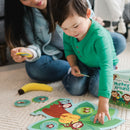 A young boy wearing a green shirt plays the game 'Monkey Around' by MindWare, interacting with colorful pieces featuring cartoon monkeys and bananas on the floor. His mother sits nearby, watching and holding a game card, with a vibrant game box visible in the background.