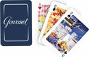 A set of Gourmet Poker Sized Playing Cards featuring artistic illustrations. The card back is a deep navy blue with the word 'Gourmet' elegantly written in white. Visible face cards include a red heart displaying drinks at a table, a black spade with a colorful fall leaf design, and another showing a still life of tableware and baked goods.