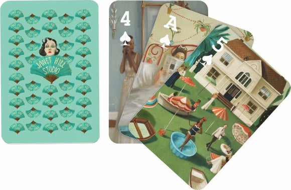 Set of Janet Hill Playing Cards by New York Puzzle Company, featuring colorful and artistic designs. The back of the cards shows a pattern of fans and the name 'JANET HILL STUDIO,' while the front displays various cards with unique artwork, including people in a garden and a house, showcasing vibrant and charming illustrations.