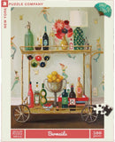 Bar Maids Janet Hill jigsaw puzzle by New York Puzzle Company, featuring a vibrant illustration of a bar cart adorned with colorful bottles, a silver bowl of lemons, and playful mermaids in the background. The puzzle contains 500 pieces and showcases an artistic design perfect for puzzle enthusiasts and art lovers alike.