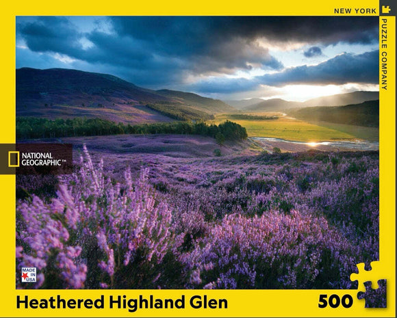 The Heathered Highland Glen Puzzle by New York Puzzle Company features a stunning landscape of rolling hills covered in vibrant purple heather flowers, under a dramatic sky filled with clouds. This 500-piece jigsaw puzzle captures the serene beauty of nature, perfect for puzzle enthusiasts and nature lovers alike.