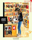 A jigsaw puzzle titled 'At the Strand', featuring an illustration of a woman browsing books in a cozy bookstore. The cover art by Jenny Kroik captures a warm, inviting atmosphere with shelves filled with colorful books. This 1000-piece puzzle is created by New York Puzzle Company and is ideal for puzzle enthusiasts and fans of literature.