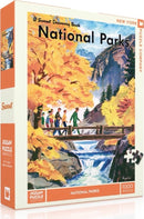 New York Puzzle Company's National Parks Jigsaw Puzzle featuring vibrant autumn scenery with a group of hikers on a bridge over a stream. The puzzle includes 1000 pieces and showcases a picturesque landscape with colorful foliage and mountains in the background.