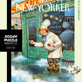 A 1000-piece jigsaw puzzle titled 'Just a Pinch' by New York Puzzle Company, featuring a whimsical illustration of a chef outside a cafe, snowflakes falling around him. The design showcases a charming winter scene with warm, inviting colors and detailed artistry.