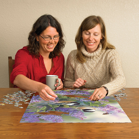 Two women engaged in assembling a colorful jigsaw puzzle titled 'Chickadees and Lilacs' by Cobble Hill, featuring a beautiful design of lilacs and birds. They are seated at a wooden table with scattered puzzle pieces and a coffee mug visible.