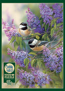 A beautifully illustrated jigsaw puzzle titled 'Chickadees and Lilacs' by Cobble Hill. The image features two chickadee birds perched on lilac branches, surrounded by vibrant purple flowers and lush green leaves. This 1000-piece puzzle comes with a poster included for reference.