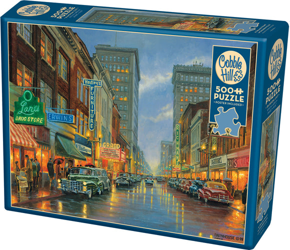 A Grand Night in Steubenville jigsaw puzzle by Cobble Hill featuring a vibrant street scene illuminated by neon signs and reflecting lights on wet pavement. The puzzle contains 500 pieces and includes a poster for reference.