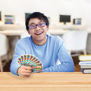 A happy young man wearing glasses holds a colorful deck of WordSpiel cards, sitting at a wooden table. The background features a bright and modern study environment with computers and stacks of books.