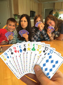 A group of four people, two children and two adults, are seated around a table playing a card game called Five Crowns. They are holding colorful cards and displaying various facial expressions of concentration and fun. The game offers an engaging family experience.