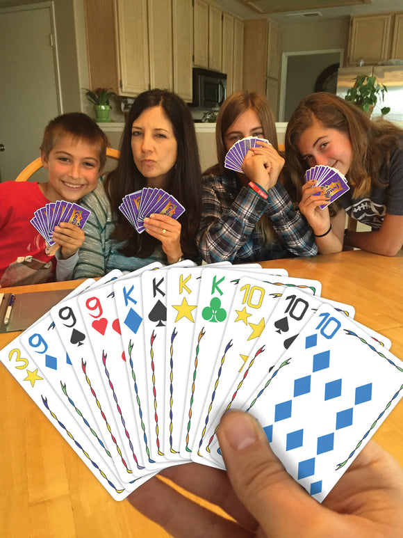 A group of four people, two children and two adults, are seated around a table playing a card game called Five Crowns. They are holding colorful cards and displaying various facial expressions of concentration and fun. The game offers an engaging family experience.