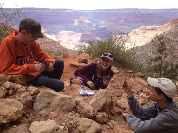 Three friends enjoy a game of Five Crowns while sitting on rocks amidst a scenic backdrop of the Grand Canyon. They are laughing and engaged in lively conversation, showcasing the game's social aspect and outdoor fun.