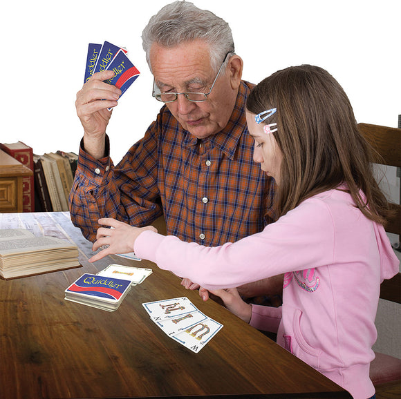 A grandfather and granddaughter playing Quiddler, a word game, at a wooden table. The grandfather holds cards while the granddaughter points at cards laid out. A book is open nearby, enhancing the cozy game atmosphere.