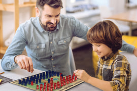 A man and a young boy are engaged in a game of Stratego at a table. The game board features red and blue pieces arranged in a strategic layout, with the pair focused and smiling as they play together, fostering a sense of competition and fun in a cozy indoor setting.