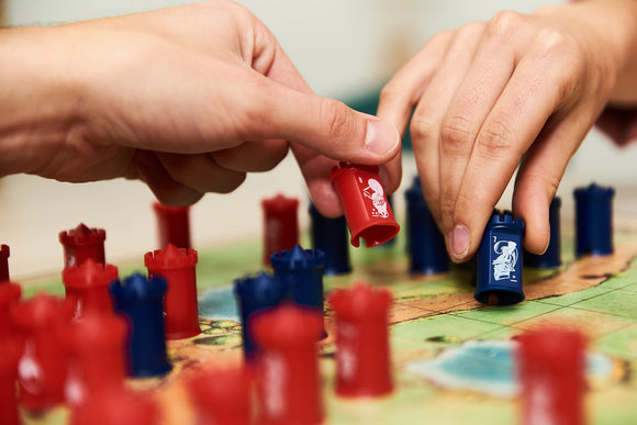 Two people playing Stratego, a strategy board game, manipulating red and blue pieces on a colorful game board. The hands are focused on a colorful battlefield as they strategize their moves, highlighted by detailed character illustrations on each piece.