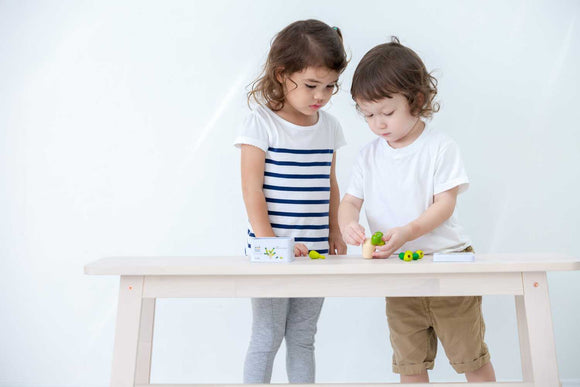 Two children play with the Balancing Cactus Mini game by Plan Toys on a wooden table. One child focuses on stacking colorful cactus pieces, while the other observes with interest. The game encourages creativity and balance in a fun, interactive way.