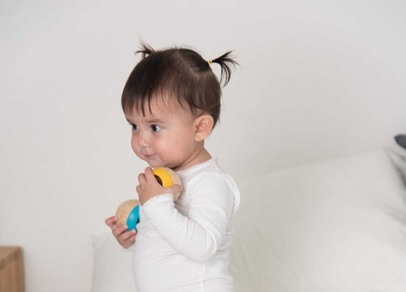 A toddler with pigtails holds colorful wooden balls, engaging playfully. The child is dressed in a white long-sleeve top, showcasing joy and curiosity in a bright setting.