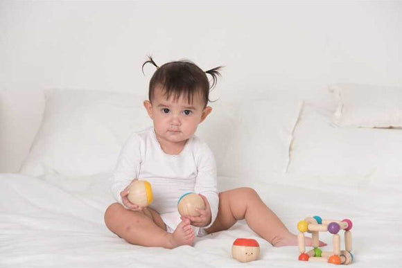 A young child sitting on a bed, engaging with colorful wooden sensory toys. The child holds two round, wooden balls with different colors, while a small block and a multi-colored connector toy are nearby.