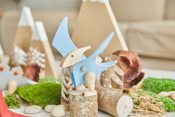 The DIY Pterodactyl kit by Plan Toys features a vibrant blue wooden pterodactyl figure set amidst a playful scene with soft green moss, wooden logs, and decorative elements resembling trees and mountains, designed for imaginative play and creativity.