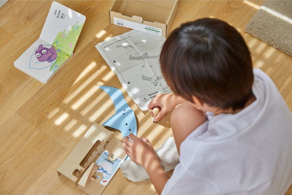 A child assembling the DIY Pterodactyl kit by Plan Toys. The scene shows the child engaged with colorful pieces, an instruction booklet, and a partially assembled model on a wooden floor, surrounded by warm sunlight.