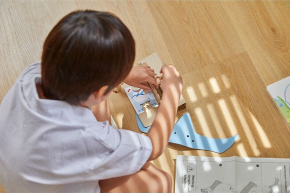 A child focuses on assembling the DIY Pterodactyl kit from Plan Toys. The scene captures the wooden pieces laid out on the floor along with a set of instructions, showcasing creativity and hands-on play.
