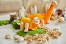 A DIY Triceratops kit by Plan Toys featuring colorful wooden components. The kit includes a bright yellow Triceratops with a hat and a playful dinosaur set on a green landscape of moss and faux dirt, surrounded by small rocks.