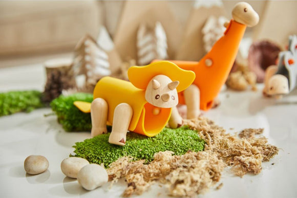A DIY Triceratops kit by Plan Toys featuring colorful wooden components. The kit includes a bright yellow Triceratops with a hat and a playful dinosaur set on a green landscape of moss and faux dirt, surrounded by small rocks.