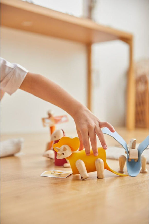 A child plays with a colorful DIY Triceratops toy from Plan Toys. The wooden dinosaur features a vibrant yellow body and small wooden limbs, engaged in imaginative play among other colorful animals. The warm, inviting environment suggests a playful atmosphere.