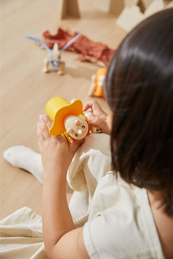 A child carefully assembles a colorful DIY Triceratops kit designed by Plan Toys. The kit features vibrant yellow, orange, and wooden elements, encouraging creativity and imaginative play.