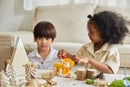 Two children engaged in a DIY Triceratops kit by Plan Toys. One child is using a tool to modify a colorful dinosaur figure, while the other looks on attentively. Surrounding them are various natural materials like wood slices, pinecones, and crafted nature elements, creating a playful, creative environment for hands-on learning.