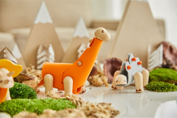 A colorful DIY Brachiosaurus kit from Plan Toys, featuring an orange dinosaur with wooden limbs and a smiling face. The scene includes a playful landscape with greenery and other dinosaur figures, showcasing creativity and imagination.