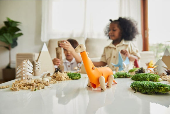 A bright orange DIY Brachiosaurus kit from Plan Toys is prominently displayed on a tabletop surrounded by natural materials and decorative elements like trees and foliage. Two children in the background are engaged in creative activities, one building a tower and the other holding a blue dinosaur shape. The scene conveys a playful and imaginative atmosphere for hands-on learning.