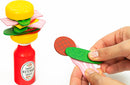 The Burger Balance game by Professor Puzzle features a colorful stacking challenge with playful food pieces. A hand is seen adding a green pickle slice on top of a stacked burger with a ketchup bottle base, showcasing vibrant colors and fun shapes designed to engage players in a balancing act.