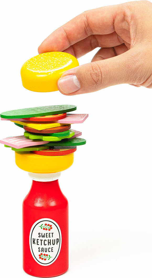 A colorful stacking game called Burger Balance by Professor Puzzle. The image shows a hand carefully placing a yellow burger bun on top of a stack of colorful burger ingredients, balanced on a bottle labeled 'Sweet Ketchup Sauce.' The vibrant pieces include lettuce, cheese, and tomato, creating a fun, engaging scene.