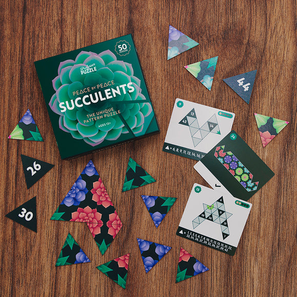 The 'Peace by Peace: Succulents' mechanical puzzle from Professor Puzzle. The image shows the colorful box featuring succulent patterns, alongside its 50 unique triangular pieces. Several puzzle pieces, some numbered, are spread out on a wooden surface, along with instruction cards displaying different puzzle patterns.
