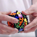 A person holding a colorful Gear Ball mechanical puzzle by Mefferts. The spherical puzzle features various geometric shapes and vibrant colors including orange, green, blue, yellow, and purple. The intricate design allows for rotation and manipulation, providing a fun and challenging experience for puzzle enthusiasts.