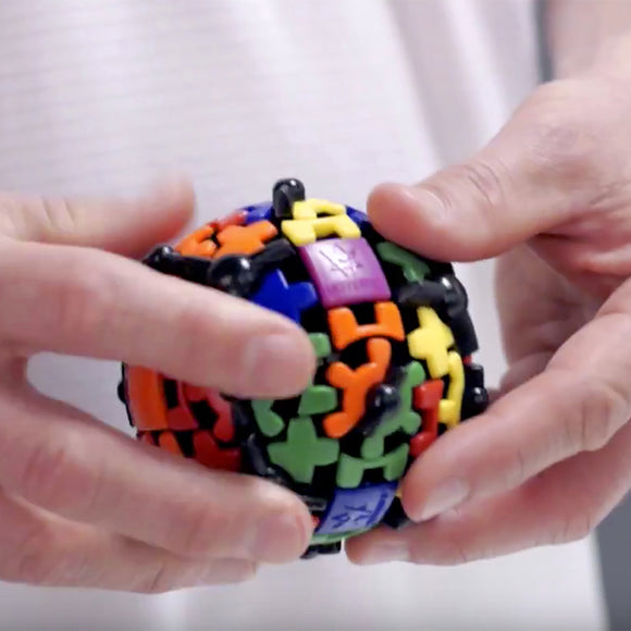 A person holding a colorful Gear Ball mechanical puzzle by Mefferts. The spherical puzzle features various geometric shapes and vibrant colors including orange, green, blue, yellow, and purple. The intricate design allows for rotation and manipulation, providing a fun and challenging experience for puzzle enthusiasts.