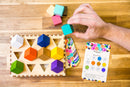 The Chroma Cube mechanical puzzle by Project Genius features a colorful arrangement of geometric blocks, with a hand positioning a teal cube. The wooden base displays assorted shaped slots for the blocks, and an instructional card with clues is visible, providing a challenging and engaging experience for puzzle enthusiasts.