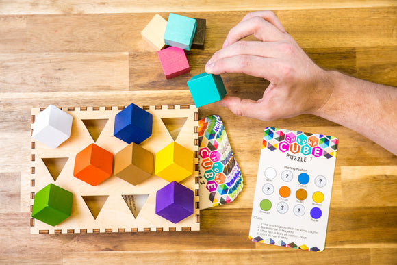 The Chroma Cube mechanical puzzle by Project Genius features a colorful arrangement of geometric blocks, with a hand positioning a teal cube. The wooden base displays assorted shaped slots for the blocks, and an instructional card with clues is visible, providing a challenging and engaging experience for puzzle enthusiasts.
