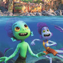 A colorful underwater scene featuring two happy sea creature characters. The character on the left has green skin, big eyes, and a wide smile, while the character on the right has purple skin and is wearing a yellow tank top and orange shorts. They are playfully swimming near rocks and colorful buildings visible in the background above the water's surface.