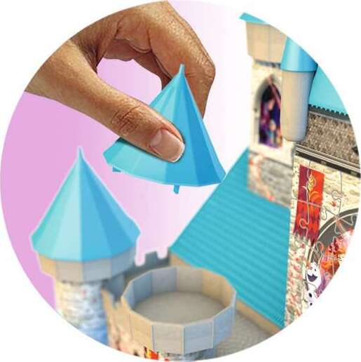 A close-up of a hand holding a blue, cone-shaped piece above a castle structure from the Ravensburger Frozen Castle jigsaw puzzle. The scene shows the beautiful, colorful details of the castle with its distinctive blue roofs and gardens.