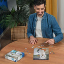 A person is sitting at a wooden table, working on the Puzzle Moments: Alpaca 200 pc jigsaw puzzle by Ravensburger. The puzzle features an adorable alpaca with a floral decoration around its neck. The completed section of the puzzle is displayed in front of them, while the remaining puzzle pieces are scattered around. A puzzle box with the same design and a green plant can be seen in the background.