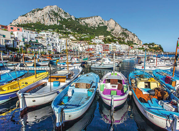 A vibrant jigsaw puzzle titled 'Colorful Marina' by Ravensburger. The image features a picturesque scene of a marina with colorful boats docked, set against a backdrop of charming coastal buildings and a striking mountainous landscape under a clear blue sky.