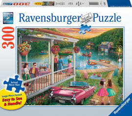 Ravensburger jigsaw puzzle titled 'Summer at the Lake' featuring a vibrant scene of people enjoying a lakeside setting with colorful flowers, boats, and a classic car. The puzzle has 300 pieces, large piece format for easy handling, and showcases a relaxed summer atmosphere.